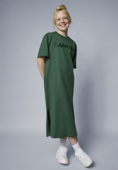 Zelena pamučna haljina s kratkim rukavima, bočnim rezovima i 'LANVIN' logotipom tamnije zelene boje. Model nosi bijele tenisice i čarape. Glatka tekstura.