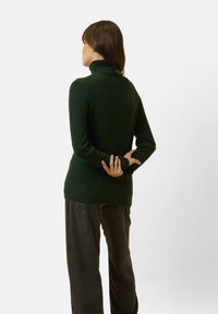 Maglione a collo alto verde scuro realizzato in un materiale morbido e texture. Il design è aderente in vita e si estende oltre i fianchi.