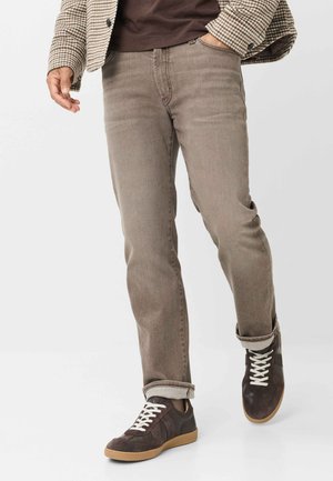 Mand gående iført beige jeans, brun ternet jakke og mørkebrune sneakers med hvide snørebånd på hvid baggrund.