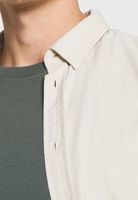 Camisa beige de botones con una textura sutil, cuello abierto, superpuesta sobre una camiseta ajustada de color verde oscuro. Costuras visibles y detalle de botones.