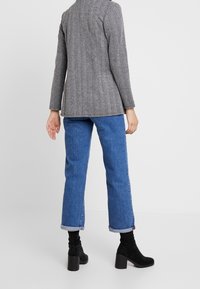 Šedý herringbone blazer s prodlouženou siluetou, kombinovaný s modrými džíny s ohrnutými manžetami a černými kotníkovými botami na podpatku.