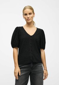 Kvinde med blondt hår sat op, iført en sort struktureret top med pufærmer og knapper, mørkegrå jeans, sølvfarvede hoop øreringe og armbånd.