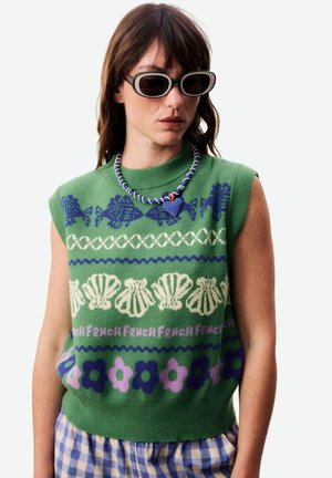Femme portant des lunettes de soleil noires, un pull sans manches vert orné de motifs de poissons bleus, coquillages, fleurs et le mot « FRENCH », ainsi qu'un collier avec un cœur bleu.