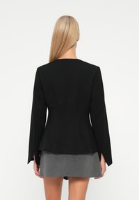 Vrouw met lang blond haar draagt een getailleerde zwarte blazer met gespleten mouwen en een korte grijze rok, staand voor een witte achtergrond.
