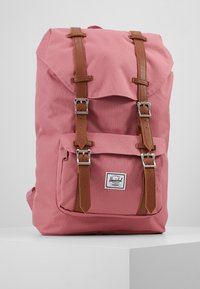 Sac à dos en toile rose avec des sangles et des boucles en cuir marron, une poche avant et une fermeture à rabat. Présente un logo de la marque sur le bas du devant.
