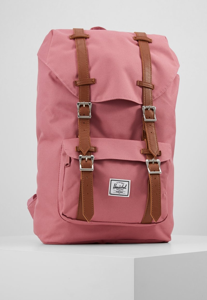 Sac à dos en toile rose avec des sangles et des boucles en cuir marron, une poche avant et une fermeture à rabat. Présente un logo de la marque sur le bas du devant.