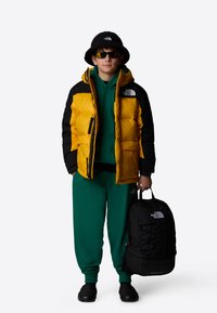 Giacca a vento gialla e nera, felpa verde con cappuccio, pantaloni della tuta verdi, cappello a secchiello nero, occhiali da sole neri e zaino nero con dettagli del logo.