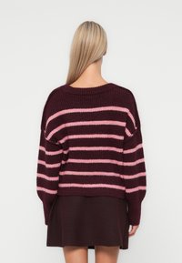 Pull en tricot rayé bordeaux et rose avec une coupe décontractée, encolure côtelée et épaules tombantes. Longueur cropped, porté sur une jupe sombre.