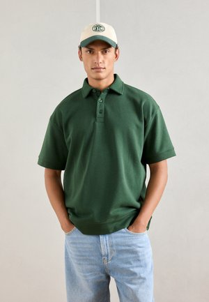 Polo shirt en coton vert avec col et trois boutons, associé à un jean en denim bleu clair. Le modèle porte une casquette beige avec des accents verts.