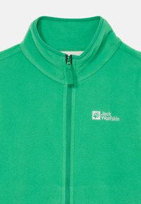 Roheline fliisjakke kaelusega ja lukuga ees. Vasakul küljel on valge "Jack Wolfskin" logo. Pehme tekstuur.