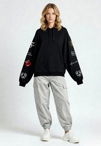 Juodas per didelis gobtuvu paslėptas megztinis su grafikos spausdinimu ant rankovių, kišene ir raišteliu. derinamas su šviesiai pilkomis krovinių kelnėmis ir baltomis sportbačiais.