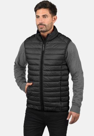 Chaleco acolchado negro con cremallera frontal, presenta un acolchado horizontal y una camiseta de manga larga gris debajo. Textura suave, diseño elegante.