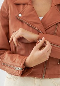 Hnědá kožená motocyklová bunda se stříbrným kováním. Obsahuje zipové manžety a stylový asymetrický přední zip. Texturovaný povrch.