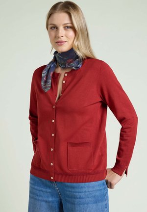 Woman wearing a red buttoned cardigan with a front pocket, blue jeans, and a dark floral neck scarf, standing with hands on hips.
