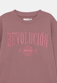 Sudadera de cuello redondo en color malva con el texto bordado en rosa "REVOLUCIÓN" y un pequeño emblema con "Senna Vingino" y los años '07 y '97.