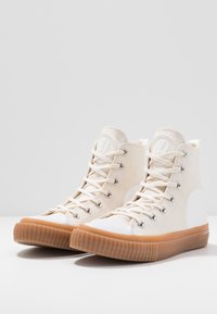 Höga sneakers gjorda av krämfärgad kanvas med vit gummikappa på tån, med silverfärgade ögla, kontrasterande tan färgad ribbad sula och logotypdetalj.