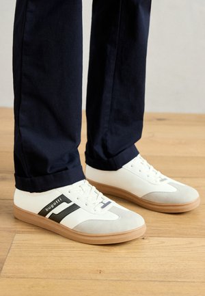 Fehér és szürke sportcipők fekete csíkokkal, bőrből készültek, gumitalppal és lapos fűzővel. Tengerészkék nadrággal viselik egy fa padlón.