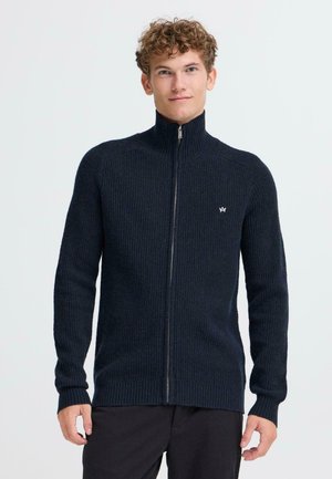 Marineblå ribbestrikket zip-up genser med høy krage, lange ermer og logo detalj på brystet, laget av teksturert strikket materiale.