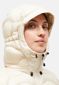 Kremfarvet dunjakke med høj krave og hætte. Har vandrette syninger, snørejusteringer og sorte detaljer på lynlåse.