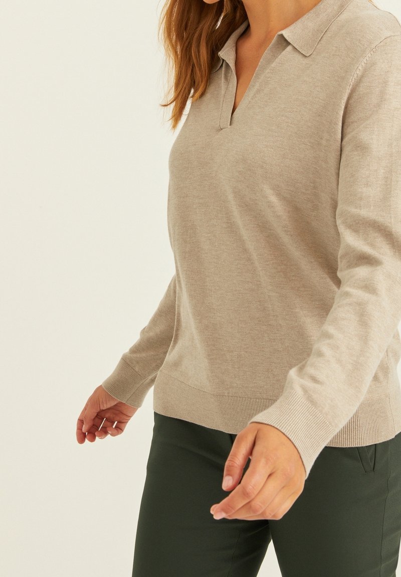 Femme portant un haut beige en maille à manches longues avec col et un pantalon vert foncé, visible du cou aux hanches sur un fond uni.
