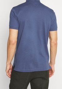 Man i kortärmad blå polo tröja sedd bakifrån, ihopparad med mörka byxor mot en enfärgad bakgrund.