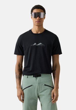 Mann trägt schwarzes T-Shirt mit Grafik, hellgrüne Hose mit Reißverschlusstaschen und große reflektierende Sonnenbrille, steht vor einfarbigem Hintergrund.