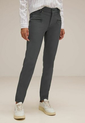 Pantalons gris ajustés avec poches avant zippées et un tissu lisse, légèrement extensible. Associés à des baskets blanches pour le contraste.