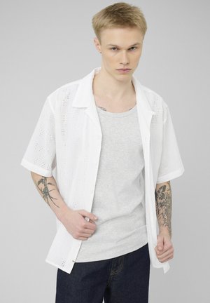 Jeune homme aux cheveux blonds courts portant une chemise blanche à manches courtes ouverte sur un débardeur gris, montrant des tatouages sur les deux avant-bras.