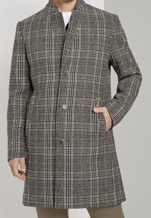 Mann trägt einen grau-schwarz karierten knielangen Mantel mit Stehkragen und Knöpfen, rechte Hand in der Tasche.