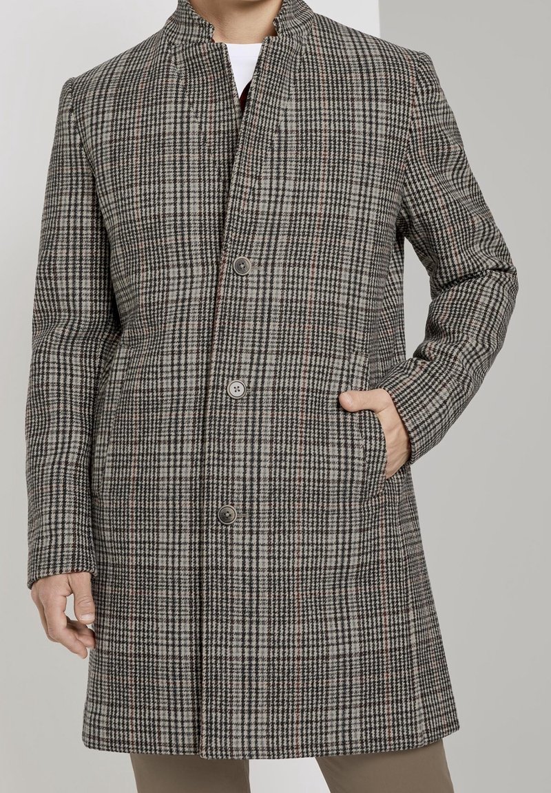 Hombre con abrigo a cuadros gris y negro hasta la rodilla, con cuello alto y botones, con la mano en el bolsillo derecho.