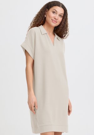 Jeune femme aux cheveux bouclés portant une robe beige courte à manches courtes, sans col, longueur genou, debout devant un fond clair uni.