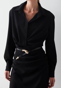 Blusa nera con colletto e maniche lunghe, caratterizzata da un design frontale attorcigliato. Indossata con una cintura nera accentuata da una testa di serpente dorata.