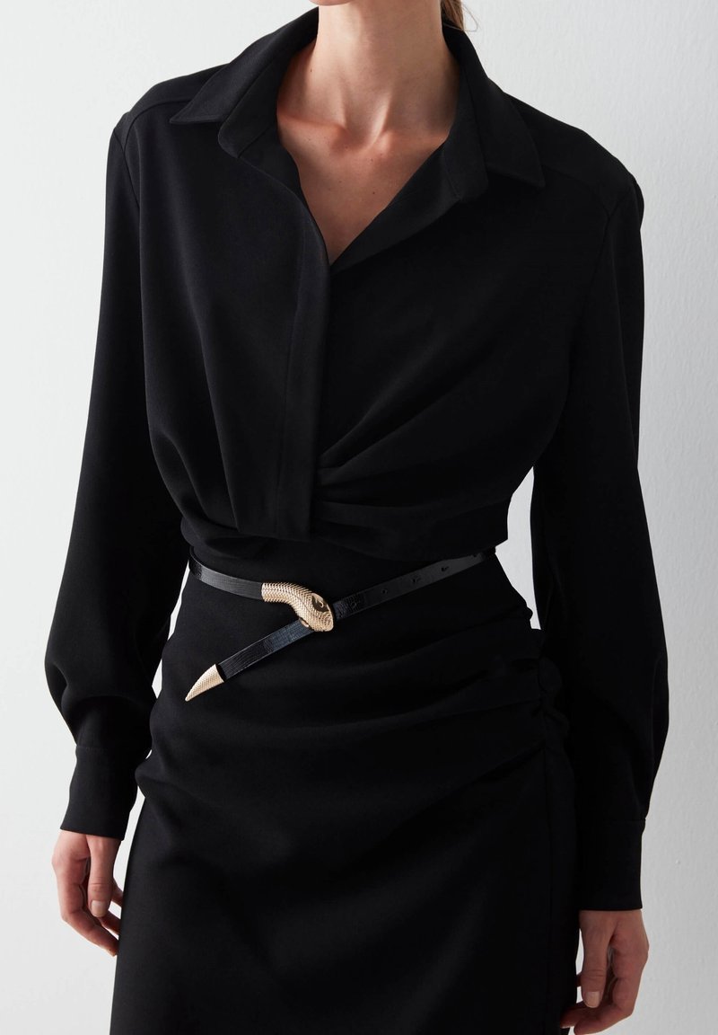 Blusa nera con colletto e maniche lunghe, caratterizzata da un design frontale attorcigliato. Indossata con una cintura nera accentuata da una testa di serpente dorata.