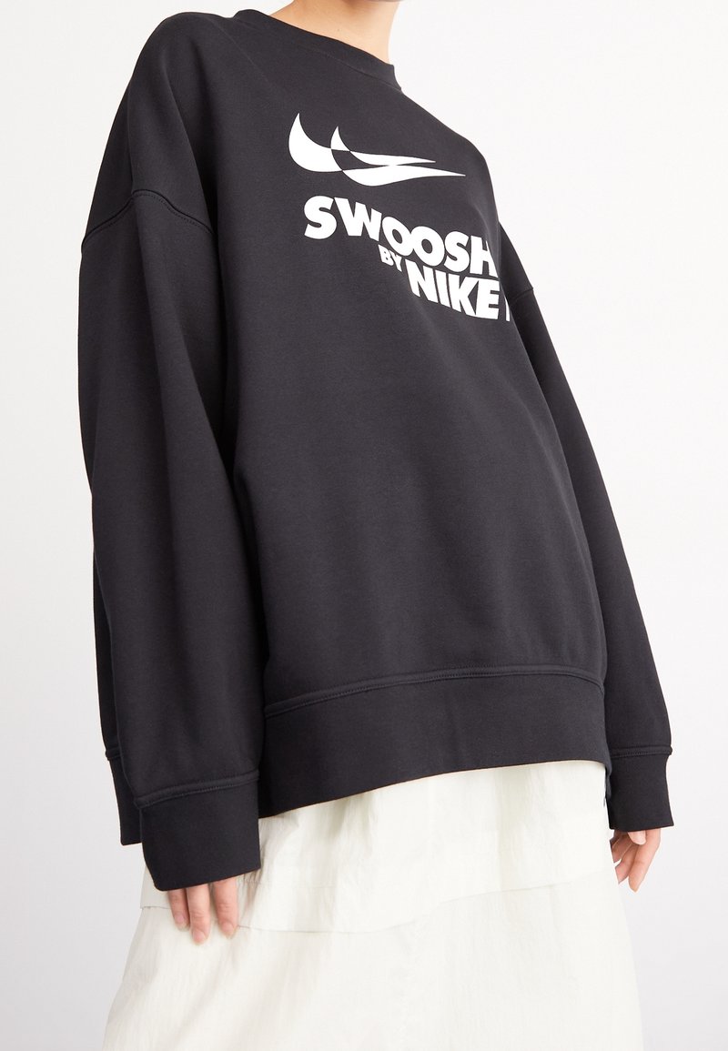 Personne portant un sweat-shirt noir surdimensionné avec le logo blanc "Swoosh by Nike" et une jupe blanche sur un fond uni.