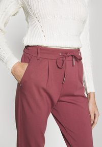 Donna che indossa un maglione bianco a maglia e pantaloni mauve a vita alta con cordoncino annodato, con la mano sinistra in tasca, su uno sfondo neutro.