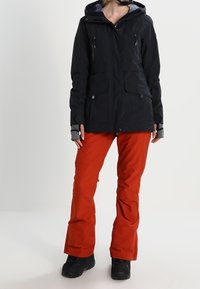 Zwarte waterdichte jas met een capuchon, voorzien van front pockets en ritsen. Gecombineerd met losse rode broeken. Zwarte laarzen maken de outfit compleet.