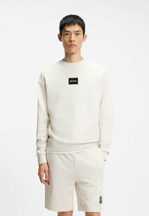 Młody mężczyzna w kremowej bluzie z logo "HUGO" i dopasowanych szortach, stojący z jedną ręką w kieszeni na białym tle.