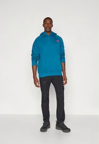 Sweatshirt com capuz azul-petróleo, com um bolso à frente e um pequeno logótipo, combinada com calças pretas e sapatos escuros. Tecido liso, corte casual.