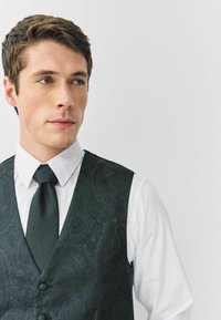 Fekete paisley mintaű mellény hat gombbal, fehér ing felett viselve, fekete nyakkendővel. Közeli felvétel, amely a felső testre összpontosít.