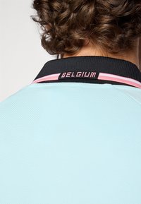Helehall sinine polosärk musta kraega, millel on roosa triip ja tagaküljel tikitud musta värvi "BELGIUM" tekst. Tekstureeritud kangas.