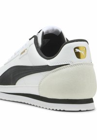 Baskets blanches avec des accents en cuir noir et un talon en daim. Présentent une rayure noire texturée et un logo doré sur le talon. Semelle en caoutchouc.