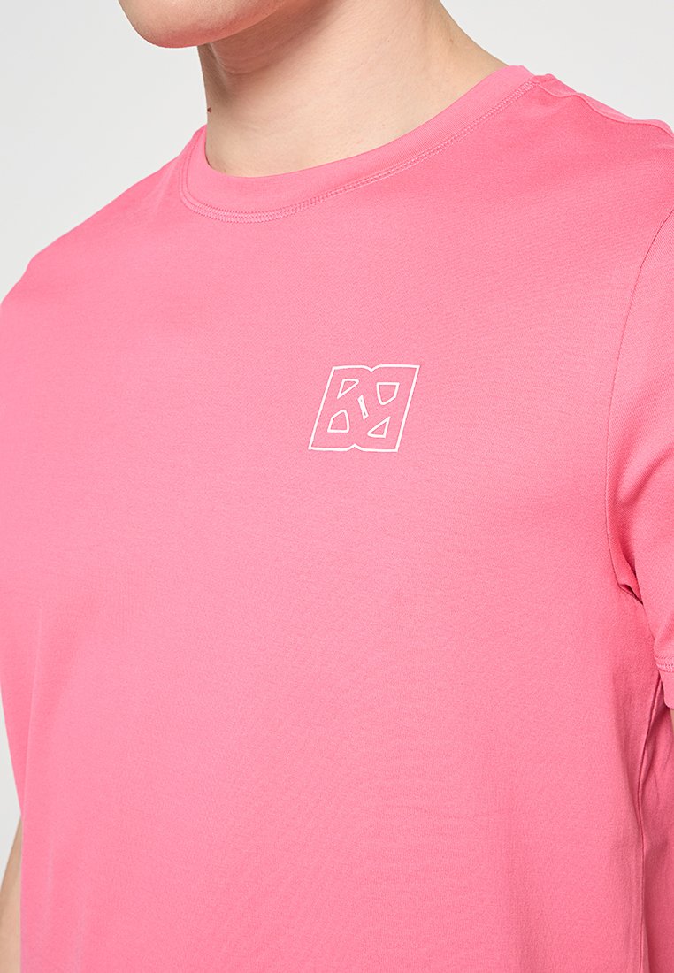 Różowa bawełniana koszulka z okrągłym dekoltem i krótkimi rękawami, z małym białym logo po lewej stronie klatki piersiowej. Gładka faktura.