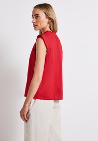 Blusa rossa senza maniche con scollatura rotonda e dettagli a ruches, realizzata in tessuto leggero, abbinata a pantaloni larghi color avorio.