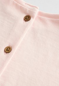 Tejido de color rosa claro con una textura suave, que presenta un escote redondo y dos botones marrones cerca de la parte superior. Diseño simple y minimalista.