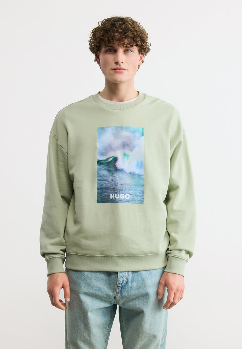 Νεαρός άνδρας με κατσαρά μαλλιά που φοράει ανοιχτοπράσινο φούτερ με μεγάλο σχέδιο ωκεάνιας κύματος και το κείμενο «HUGO», συνδυασμένο με ανοιχτό μπλε τζιν.