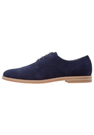 Zapato de ante azul marino con puntera redondeada, diseño de cordones con cinco ojales y suela de goma marrón claro. Detalle de costuras minimalistas.