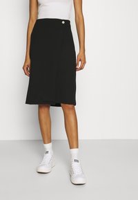 Black knee-length skirt with a wrap design, adorned with a single button at the waist. Paired with white sneakers and ankle socks.