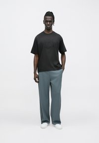 Μαύρο κοντομάνικο μπλουζάκι με ανάγλυφο λογότυπο στο στήθος, συνδυασμένο με φαρδιά, ανοιχτό γκρι παντελόνια και λευκά αθλητικά παπούτσια.