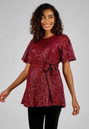 Maglia per maternità in profondo bordeaux con maniche corte a farfalla, vita con lacci e decorazioni in paillettes, abbinata a pantaloni neri.