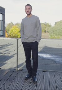 Grå långärmad sweatshirt med framficka, kombinerad med svarta byxor och svarta stövlar, stående på ett tak med trall.